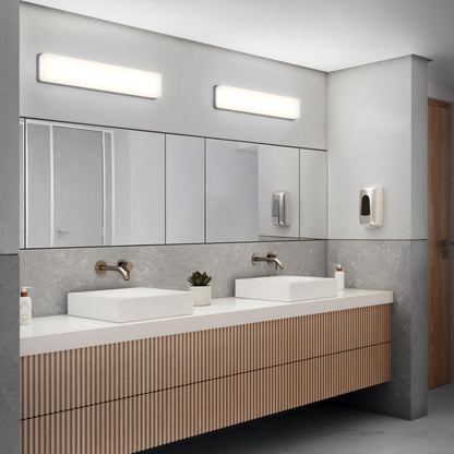 Modern public restroom with two rectangular sinks, wall-mounted faucets, large mirrors, soap dispensers, a small potted plant, and bright Aster 27in Energy Star LED Vanity Lights above the mirrors. The design is clean and minimalist.