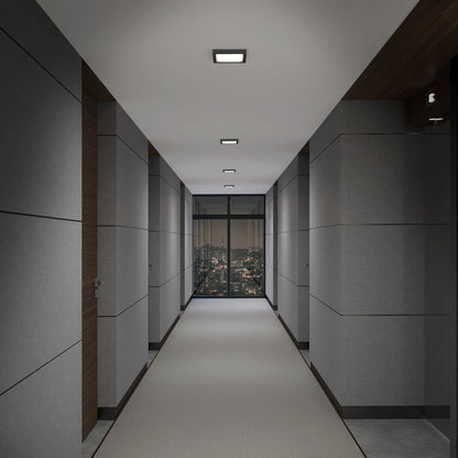 A modern hallway with grey walls features Skylight Pro Square 7in 3K LED Flush Mount ceiling lights, creating a dim, sleek look. The carpeted floor and minimalist design lead to a large window with a night city view.