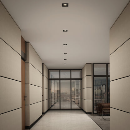 A modern hallway with beige paneled walls features Skyview 4in Regressed Square, 5CCT LED pot lights and floor-to-ceiling glass doors overlooking a city skyline. A brown chair sits in the corner near the windows.