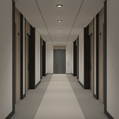A modern hallway with light walls and carpet, featuring several dark-framed doors and illuminated by 5" LED Skylights with 3 colour trim rings, leads to closed double elevator doors at the end.