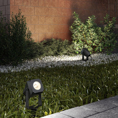 Two Verner Plug-in Landscape Single CCT LED spotlights from the 4-pack cast light on green bushes and a brick wall at night, highlighting grass and white pebbles in both the foreground and background.