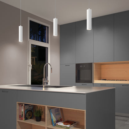 A modern kitchen with gray cabinets, a spacious island with an open bookshelf, a built-in oven, sleek faucet, and three Damian LED Cylinder Pendants illuminate the space beside a window overlooking trees.