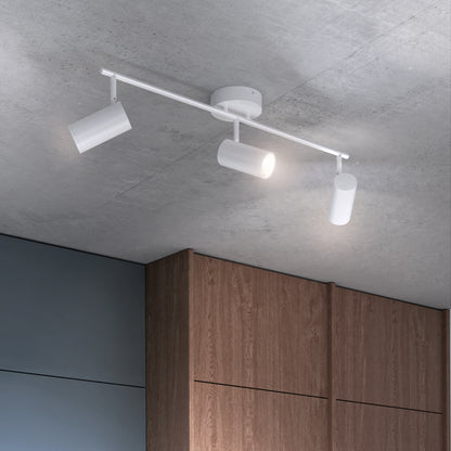 The Alpha LED Track 3-Light, a modern ceiling fixture with three adjustable white cylindrical spotlights on a metal bar, is installed on a gray concrete ceiling above wooden cabinets. One spotlight is illuminated.