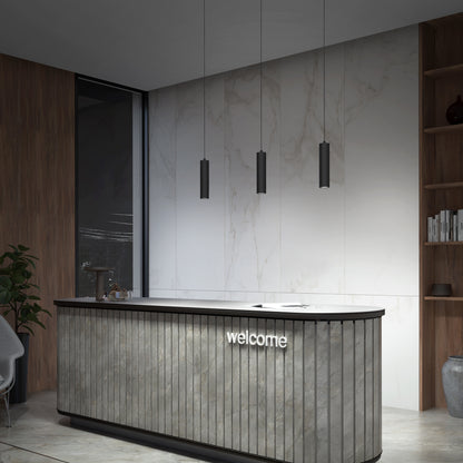 A modern reception desk with a welcome sign sits against a marble wall. Three Damian LED Cylinder Pendants hang above, while nearby bookshelves and a plant enhance the sleek, minimalist atmosphere.