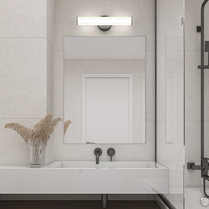 A modern bathroom with a large mirror above a white sink, matte black faucet fixtures, and the sleek Kalio Indoor LED Wall Sconce 18" in matte black. A vase with dried pampas grass rests on the counter near the glass shower enclosure.