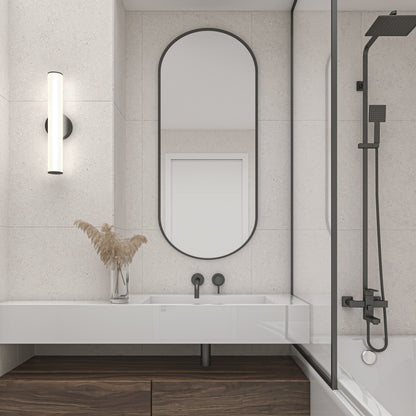 Modern bathroom with an oval mirror, wall-mounted faucet, Kalio Indoor LED Wall Sconce 18", glass shower divider, black showerhead, and a white sink on a wooden vanity. Dried grasses in a vase add decor to the counter.