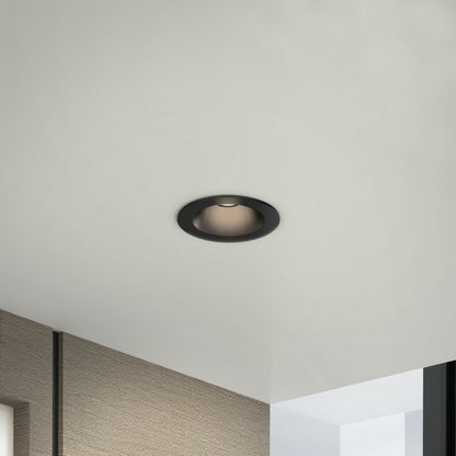 A Skyview 4in Regressed Round, 5CCT LED pot light with black trim is recessed in a white ceiling above a room featuring textured beige walls and a glass panel.