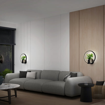 A modern living room with a gray sofa, black side table, and Halo Planter LED wall sconces displaying potted plants. Light wood and white panel walls offer contrast, while a window showcases lush greenery outside.