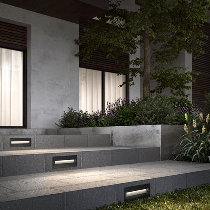 Modern outdoor steps illuminated by Dark Sky Step Light Pro- Black 12in, leading to a house with large windows, surrounded by landscaped plants and a small tree at night.
