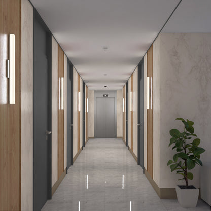 A clean, modern hallway features light marble floors and walls, wood paneling, gray doors, and bright Mylo LED Vanity Light 24INs. At the end stands a closed silver elevator, with a green potted plant on the right side.