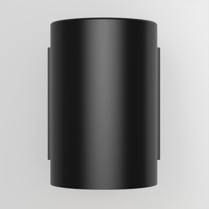 The Walton Indoor/Outdoor LED Wall Light, featuring a smooth matte black cylindrical design, is mounted on a plain white wall and showcases a minimalist, modern aesthetic when viewed from the front.