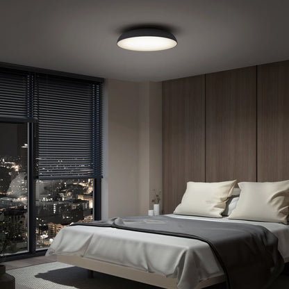 A modern bedroom at night features a neatly made bed with white and gray bedding, wooden wall paneling, large windows with dark blinds overlooking the city, and a glowing Flint LED Flush Mount ceiling light.