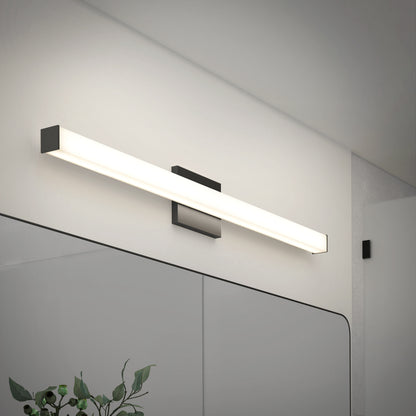 The Mylo LED Vanity Light 36IN, a modern rectangular wall-mounted fixture, is installed above a bathroom mirror on a white wall, casting bright, even illumination. Green plant leaves appear in the bottom left corner.