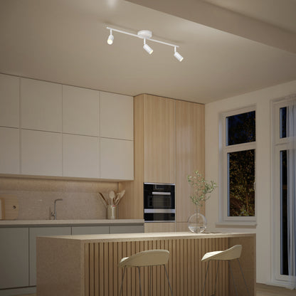 Modern kitchen with light wood and white cabinets, a built-in oven, minimalist island and two stools, vase with branches, and a 32" Rowen 3L tracklight overhead. Large windows offer nighttime views.