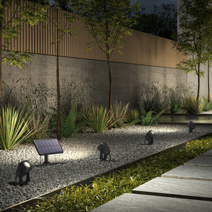 At night, a landscaped garden is lit by Verner - Solar Landscape Single CCT LED 4-pack spotlights, highlighting plants and gravel along a modern concrete and wood fence, while stone pavers form a walkway to the right.