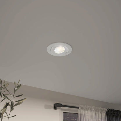 The Skyview 4in Gimbal Round, 5CCT LED is switched on in a modern room with white walls, sheer-curtained windows, and a green plant. This recessed light offers adjustable color temperature and is JA8‑2022 compliant.