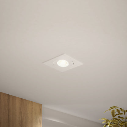 A Skyview 4in Gimbal Square, 5CCT LED recessed light glows on a smooth white ceiling, with part of a wooden wall and a leafy green plant visible, highlighting stylish contemporary indoor outdoor lighting.
