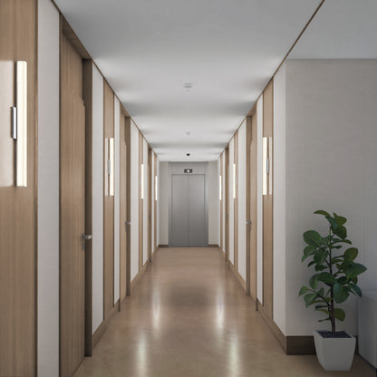 A modern hallway features Mylo LED Vanity Light 24IN fixtures on the walls, wooden doors lining both sides, a potted plant to the right, and a closed elevator at the corridor’s end.