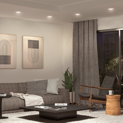 A modern living room with a gray sofa, wooden chair, coffee table, potted plant, abstract wall art, gray curtains, and a large window at night is brightly lit by the Skylight Pro 5 3K LED Flat Panel Light in the recessed ceiling.