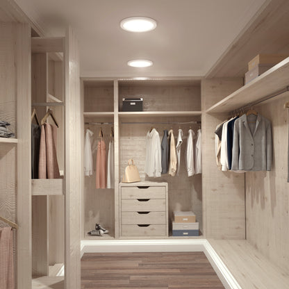 A modern walk-in closet with wooden shelves, drawers, and hanging rods displays clothes and a handbag. The space features wooden floors and is illuminated by the Skylight Pro 12 3K LED Flat Panel Light.