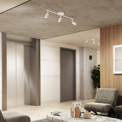 Modern living room with neutral tones, a 32" Rowen 3L tracklight on the ceiling, round coffee table, cushioned chairs, plant in the corner, and framed artwork on a wooden accent wall. The space is bright and minimalist.