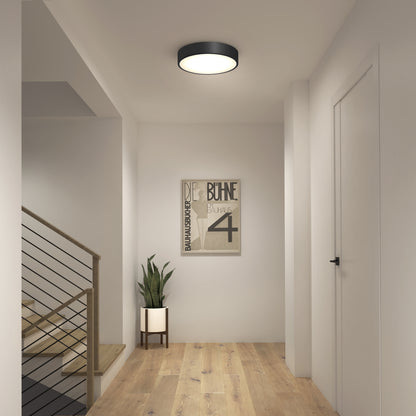 A modern hallway with light wood flooring, white walls, a Concerto LED Flush Mount ceiling light, a potted plant on a stand, a framed minimalist poster, and a staircase with a black metal railing.