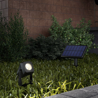 A Verner - Solar Landscape Single CCT LED 4-pack spotlight is installed on a grassy lawn by a sidewalk, with its solar panel nearby. Lush green shrubs and a brick wall form the background.