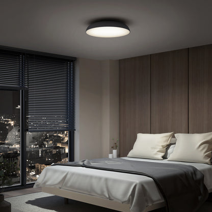 A modern bedroom at night features a large window with city lights, a neatly made bed with white and gray bedding, wood-paneled walls, and the Flint LED Flush Mount brightening the room from the ceiling.