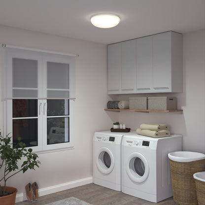 A modern laundry room with a washer and dryer, shelves and cabinets above, towels and baskets on top, a nearby laundry basket, plant, shoes, window with a blind, and bright lighting from the Knox LED 13In Energy Star Flush Mount.