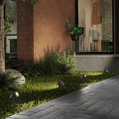 A modern outdoor garden features Verner - Plug-in Landscape Single CCT LED 4-pack lights illuminating greenery beside a brick wall and large window, with a gray stone pathway guiding past the landscaped area.