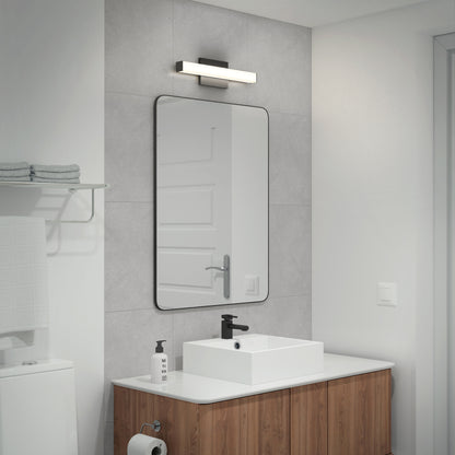 A modern bathroom features gray-tiled walls, a rectangular mirror above a white sink with wooden vanity, a soap dispenser, and the Mylo LED Vanity Light 16IN. Towels are neatly stacked on a shelf next to the toilet.