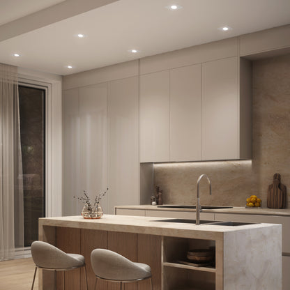 Modern kitchen with beige cabinets and marble counters features an island, two bar stools, minimalist decor, and Skyview 4in Gimbal Square 5CCT LED lights. Large window with sheer curtains ensures a bright, welcoming atmosphere.