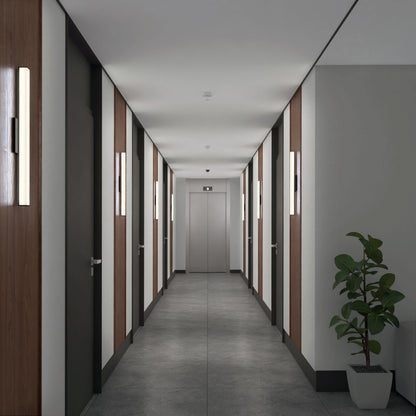 A modern hallway with gray floors, white and wood-paneled walls, and doors on both sides leads to a closed elevator. In the right corner sits a green potted plant. Lighting is provided by the Mylo LED Vanity Light 24IN.