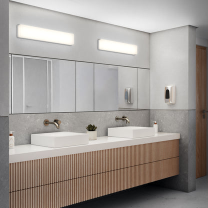 A modern public restroom features two rectangular sinks, wall-mounted faucets, mirrors, and bright Aster 27in Energy Star LED Vanity Lights overhead. The wood slat vanity holds a small plant, with soap dispensers mounted on the wall.