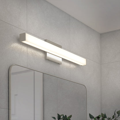 The Mylo LED Vanity Light 24IN, a modern rectangular wall-mounted fixture, is installed above a large frameless mirror on a light gray tiled bathroom wall. Green plant leaves appear at the bottom edge of the mirror.