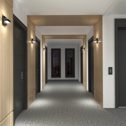 A modern, well-lit hallway with gray carpet, wooden panels, four black elevator doors, and two large windows features Mezzo LED Indoor/Outdoor Wall Sconce Lights illuminating the space.