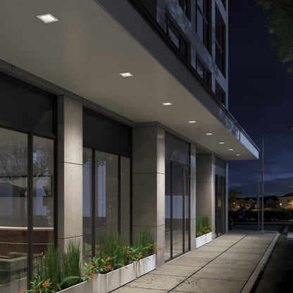 A modern building entrance at night is beautifully illuminated by Skyview 4in Regressed Square, 5CCT LED pot lights, enhancing large glass windows and planters with stylish, adjustable indoor-outdoor lighting.