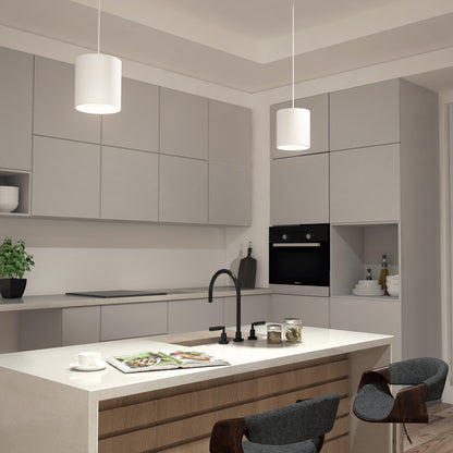 Modern kitchen with light gray cabinets, white countertops, a black faucet, built-in oven, barstools, a plant, and an island counter displaying an open magazine and cup. Features two Beam Pro 8 Integrated LED 2-IN-1 Ceiling Mount Pendants.