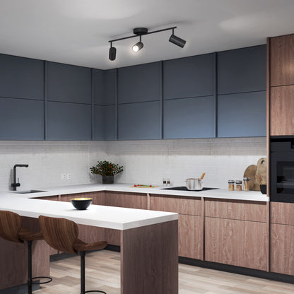 Modern kitchen featuring blue upper cabinets, light wood lower cabinets, white countertops, and an island with two wooden bar stools. The space is illuminated by the Alpha LED Track 3-Light fixture. Visible: black faucet, built-in stove, bowl.