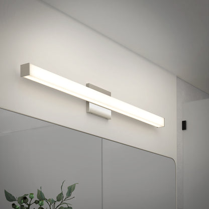 The Mylo LED Vanity Light 36IN, a modern rectangular wall-mounted fixture, is installed above a bathroom mirror, casting a bright, even glow across the white wall and mirror while green plant leaves appear in the foreground.