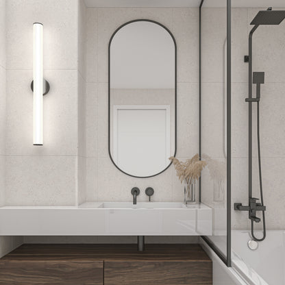 Minimalist bathroom with a large oval mirror above a white sink, sleek black fixtures, a 32" Kalio Indoor LED Wall Sconce, a glass shower with black hardware, and a vase of dried grasses on the counter.