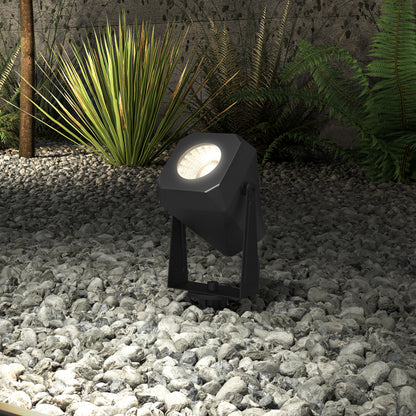 A Verner - Solar Landscape Single CCT LED spotlight from the 4-pack is placed on gray rocks, casting light on green plants and enhancing the textured stone wall in a landscaped garden.