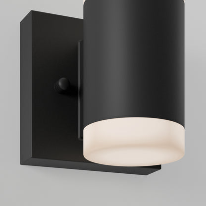 A close-up of the Neo LED Indoor/Outdoor Wall Light Stainless features a modern black cylindrical fixture on a square base, emitting a soft white glow against a light gray wall.