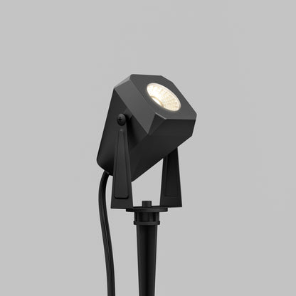 The Verner Plug-in Landscape Single CCT LED 4-pack features black, geometric outdoor spotlights on spikes for ground installation. Weather-resistant with visible power cables, they're perfect for pathway and landscape lighting. Plain gray background shown.